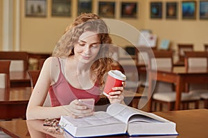 Slim good looking young student sitting in local library, holding papercup of coffee and smartphone, looking at mobile screen,