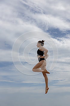 Slim gymnast woman jumping over cloudy blue sky. Young Caucasian woman wearing black sportswear. Fitness, wellness concept.