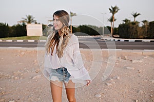 Slim long-haired girl in vintage white blouse walking on the sand with exotic palm trees on background. Charming young