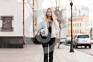 Slim pretty young woman model with long blonde hair in a fashionable white jacket in stylish black jeans in a trendy leather bag