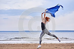 Slim red-haired middle-aged woman in sports form is engaged in dancing with a blue scarf. A woman is engaged on the sandy shore of