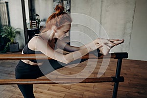 Slim redhaired woman stretching on ballet barre in classroom