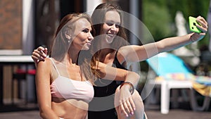 Slim selfish Caucasian women taking selfie on smartphone sitting at luxurious resort in sunshine. Portrait of two