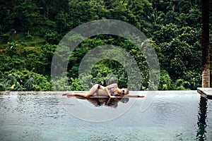Slim brunette woman in swimsuit relaxing on edge tropical infinity pool in jungle. Palms around and crystal clean
