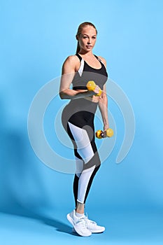 Slim sporty beautiful blonde woman doing exercises with small dumbbells isolated over blue background