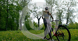 Slim sporty woman is walking in forest with bicycle, sitting on it and riding, back view