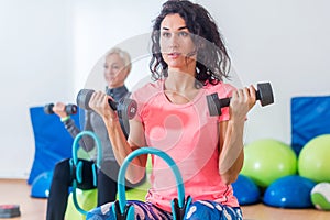 Slim sporty women training sitting on exercise balls holding dumbbells and squeezing Pilates Ring between their legs