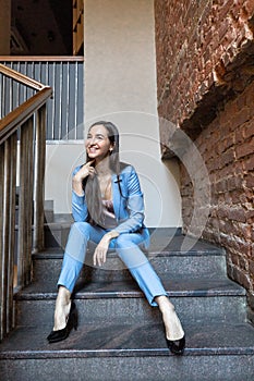 Slim stylish girl with long hair wearing blue suit sitting on the stairs. Ladies fashion and footwear in summer city.