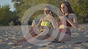 Slim tanned women drinking healthful juice as sitting on beach mat at sunset. Side view portrait of beautiful young