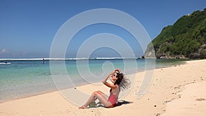 Slim tanned young woman in red bikini come and sitting on sand seashore and drinking coconut juice, enjoying wild beach