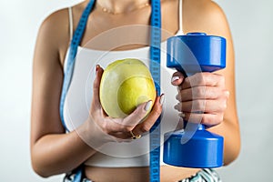 Slim unrecognizable woman with tape measure holding blue dumbbells and apple.