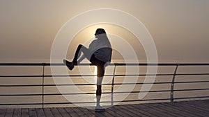 Slim woman with long legs doing stretching on sunrise by the seaside, perfect body. Stretches her leg above head
