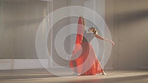 Slim woman in long scarlet red skirt and pointes jumping and bending in slow motion in backlit fog. Wide shot of