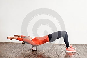 Slim woman in sport tight pants doing exercise with foam roller massager on floor, relaxing and stretching spine muscles