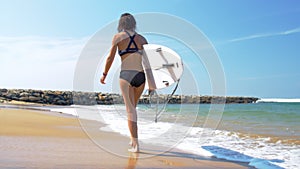 Slim woman in sports bikini with surfboard walks along shore