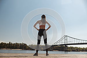 Slim woman in sportswear with hands on weist stands on embankment