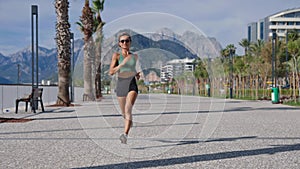 Slim young blonde woman jogging on promenade, city buildings on background. Sporty sexy woman running, exercising