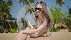 Slim young woman applying waterproof sunscreen on legs at tropical beach, palm trees. Smiling girl puts sun block on