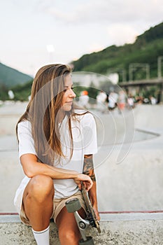 Slim young woman with long blonde hair in light sports clothes sitting with longboard in the outdoor skatepark at sunset