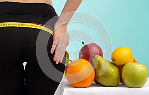 Slim young woman measuring her buttocks with a tape measure. In focus fruits for weight loss - apple, orange and pear.