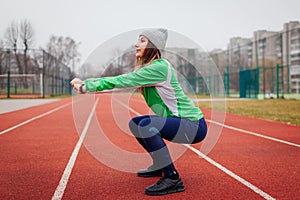 Slim young woman in sportswear doing squats sit-ups outdoors on autumn sportsground. Workout on track