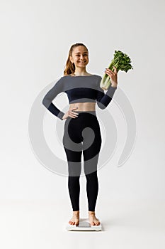 Slimming, young smiling woman stands on the scales with vegetables in her hands on a white background, vertically
