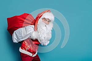 slinking santa claus carrying huge bag of gifts on blue background while looking at camera.