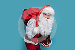 slinking santa claus carrying huge bag of presents holding gift box while looking at camera.