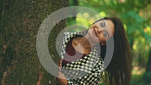 Slow mo. Happy smiling caucasian girl is showing up from tree caudex, isolated, on blurred park background. Close up