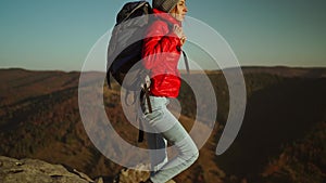 slow motion adventurous woman hiker in red jacket with backpack sits on cliff top at sunset above the woods, gets ready