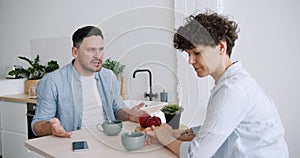 Slow motion of angry man yelling at unhappy wife sitting in kitchen at table