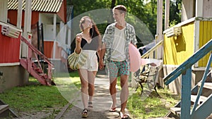 Slow motion. A beautiful couple goes to the beach on a sunny summer day, walking outdoors between the wooden houses of