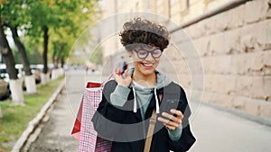 Slow motion of beautiful woman using smartphone looking at screen and walking outdoors in city holding shopping bags