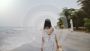 Slow motion camera follows young beautiful happy tourist woman walking along idyllic tropical ocean beach with straw hat