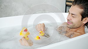 Slow motion of cheerful young man playing with toy ducks in bathtub alone