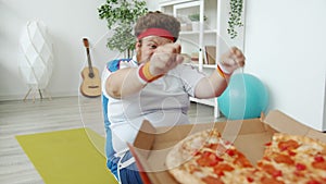 Slow motion of chubby sportsman doing press-ups and stretching hands to pizza training alone at home