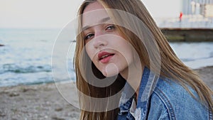 Slow motion close-up portrait of cute girl who smiles and looks at camera, sitting on beach on warm summer evening.