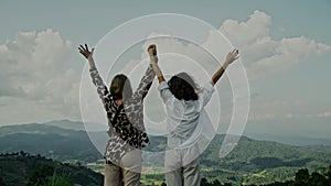 Slow motion, close-up of two young women standing on the edge of a cliff and raising their hands up against a mountain