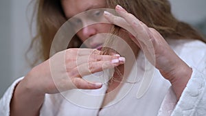 Slow motion. Close-up. A woman in a white bathrobe looks at the hair tips, which she holds in her hand, while standing