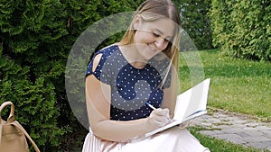 . Slow motion footage of beautiful young woman sitting in park on grass and writing a diary