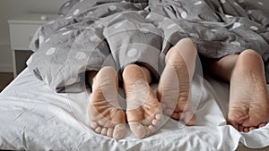 Slow motion footage of a couple's feet peeking out from under the blanket as they snuggle and relax in bed.