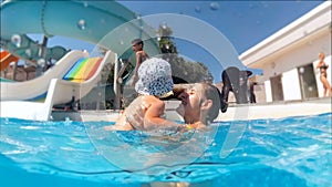 Slow motion footage of happy smiling young mother playing and throwing up her toddler boy in swimming pool at aquapark