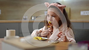 Slow motion. A girl, in a bathrobe and a pink cosmetic hair band with cat ears, sits at the table and eats pancake with