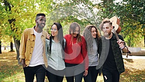 Slow motion of group of young men and women hipsters walking in park together with guitar laughing and talking. Youth