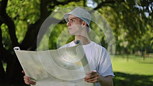 Slow motion. A guy in a sun hat holds an unfolded map in his hands and looks around while standing on a sunny summer day