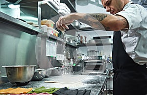 Slow motion. Handsome young chef with black tattoos on his arms pouring flour on kitchen table before making pasta