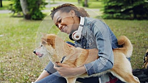 Slow motion of happy mixed race girl patting beautiful domestic dog sitting on lawn in park while glad animal is licking