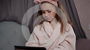 Slow motion. A little girl sits in a beautiful bedroom on a bed in a bathrobe with a pink hair band on her head and