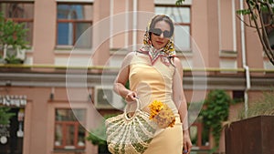 Slow motion. Low Angle Shot. A woman in a yellow dress, headscarf and sunglasses walks towards the camera, carrying a