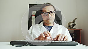 Slow motion a man in a white coat and glasses is typing on the keyboard and talking. Scientific activity of a doctor in